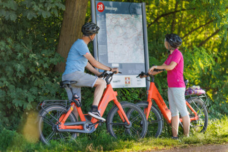 Zwei Radfahrer betrachten eine Karte an einer Infotafel mit der Aufschrift „Rheinorange“ entlang einer Radroute im Grünen.