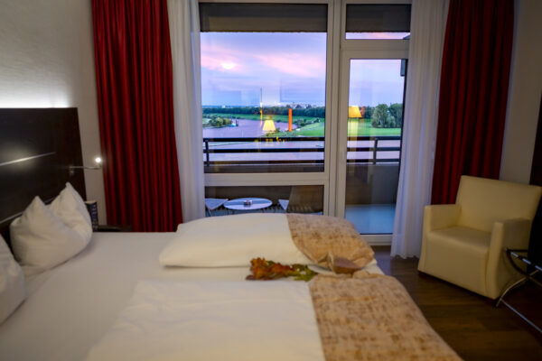Gemütliches Hotelzimmer mit Doppelbett, Sessel und Balkon mit Blick auf den Fluss bei Sonnenuntergang