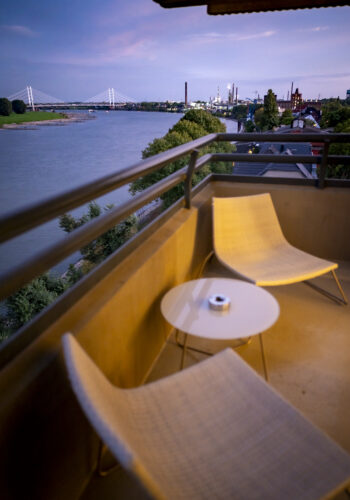 Gemütlicher Balkon im Hotel Rheingarten mit zwei Stühlen und Tisch, Blick auf den Rhein und die Brücke bei Sonnenuntergang.
