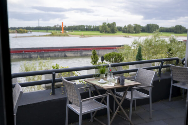 Blick von der Hotelterrasse des Hotel Rheingarten in Duisburg auf den Rhein mit vorbeifahrendem Schiff und grüner Uferlandschaft.