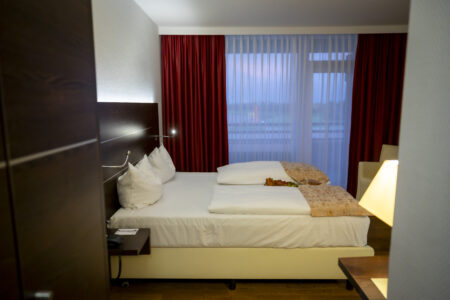Ein gemütliches Hotelzimmer im Hotel Rheingarten in Duisburg mit Doppelbett, roten Vorhängen und Blick auf den Balkon bei Abendstimmung.
