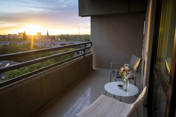 Balkon im Hotel Rheingarten in Duisburg mit Sitzgelegenheiten und Blumen auf dem Tisch, Blick auf die Stadt und den Sonnenuntergang.