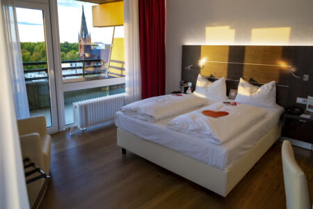 Modern eingerichtetes Doppelzimmer im Hotel Rheingarten in Duisburg mit großem Fenster und Balkon, Blick auf die Kirche und grüne Umgebung.