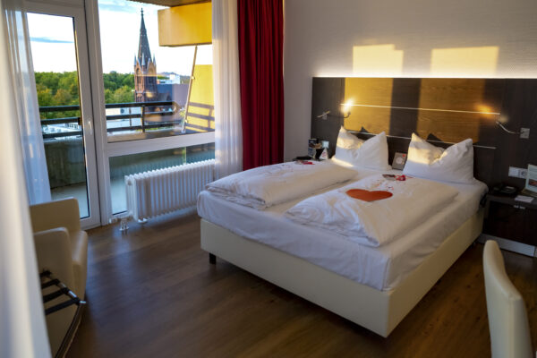 Modern eingerichtetes Doppelzimmer im Hotel Rheingarten in Duisburg mit großem Fenster und Balkon, Blick auf die Kirche und grüne Umgebung.