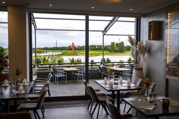 Restaurant im Hotel Rheingarten in Duisburg mit gedeckten Tischen und großem Panoramafenster, Blick auf den Rhein und die Uferwiesen.