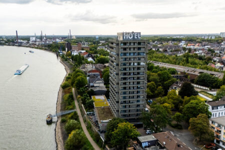Luftaufnahme des Hotel Rheingarten in Duisburg am Rheinufer mit Blick auf das Hochhaus, die angrenzende Stadt und vorbeifahrende Frachtschiffe.