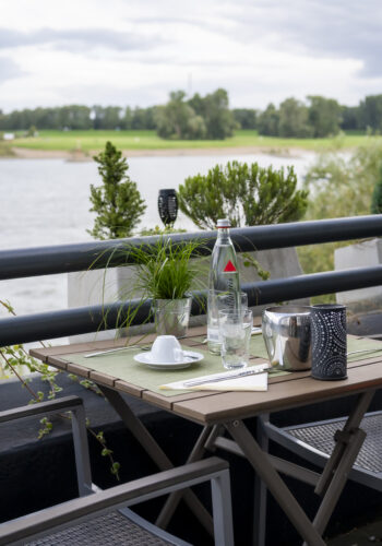 Terrasse des Hotel Rheingarten in Duisburg mit gedecktem Tisch und Sitzgelegenheiten, direkt am Rhein mit Blick auf die Uferwiesen.