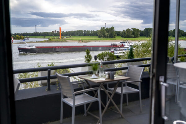 Sitzbereich auf der Terrasse des Hotel Rheingarten mit Blick auf den Rhein und vorbeifahrende Schiffe bei leicht bewölktem Himmel.