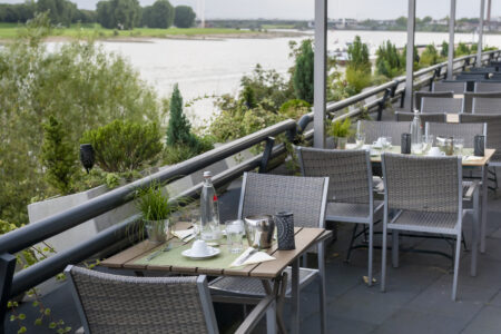 Gedeckte Tische auf der Terrasse des Hotelrestaurants mit Blick auf den Rhein und die grüne Uferlandschaft in Duisburg.