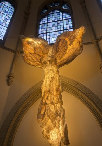 Detailaufnahme der leuchtenden Engel-Skulptur im Kolumbarium Duisburg vor bunten Kirchenfenstern.