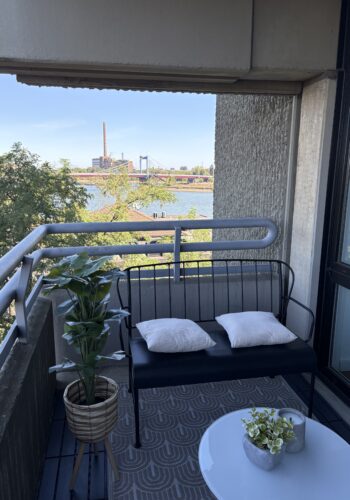 Balkon mit gepolsterter Sitzbank, rundem Tisch, Pflanze und weitem Blick über das Wasser