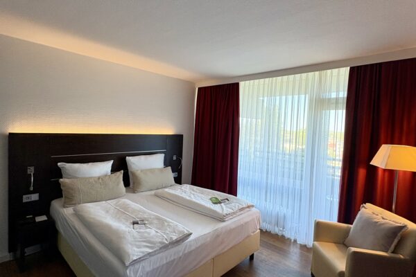 Modernes Schlafzimmer mit Doppelbett, Sessel, roter Gardine und großer Fensterfront